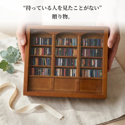 リネンの上で、両手がフタを閉じたミニチュア本棚を差し出している。そばにユーカリの小枝と無地のリボン。