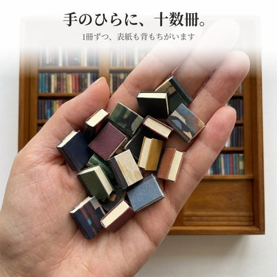 大人の手のひらにのった約20冊の小さな本。厚みの違いと、切り口のクリーム色のページがよく見える。奥にはぼけた本棚。