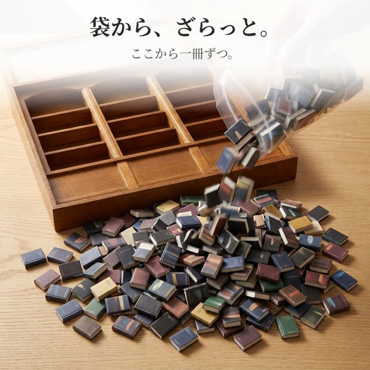 透明な袋を傾けて、小さな本が机の上にざらっと流れ出る瞬間。手前には何百冊もの山ができ、奥には空の本棚が開いている。