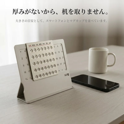 デスクに置いた薄い金属製の万年カレンダー。大きさの目安として、となりにスマートフォンとマグカップを並べている。