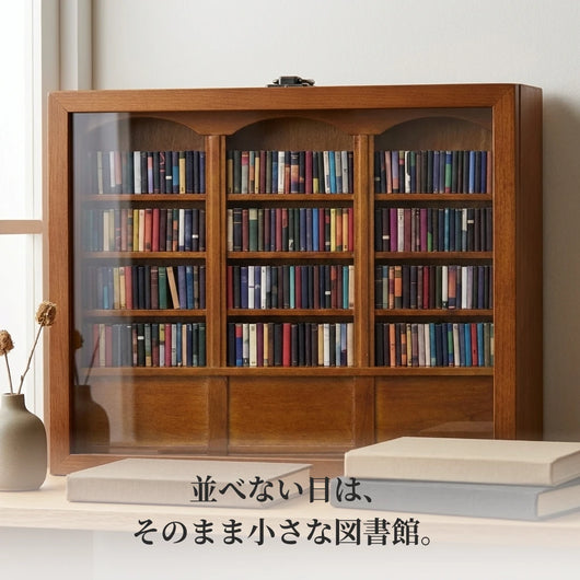 低い棚の上に、フタを閉じて立てたミニチュア本棚。無地の本と一輪挿しが添えられ、パネル越しに小さな蔵書が見える。