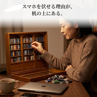 夜の机で、ランプの明かりのもと、女性が小さな本を1冊ずつ棚に並べている。ノートパソコンは閉じ、スマホは伏せてある。