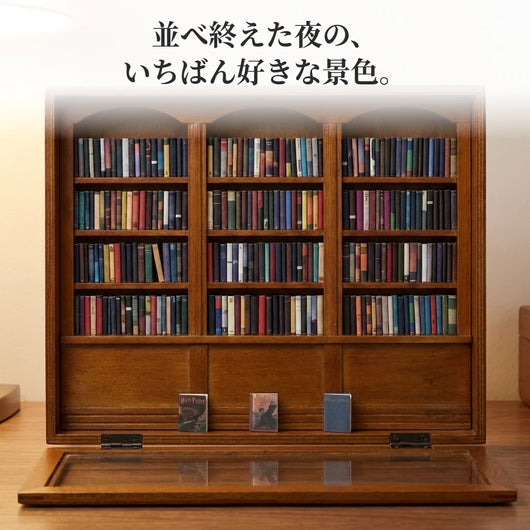 フタを手前に倒して自立させたミニチュア本棚。12区画すべてに小さな本が背を向けて並び、下の前棚には表紙を見せた本が3冊立っている。