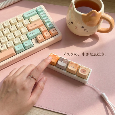 ピンクのデスクマット、パステルキーボード、猫マグの横で猫キーホルダーを押す手。「デスクの、小さな息抜き。」