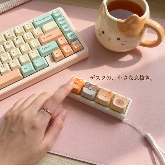 ピンクのデスクマット、パステルキーボード、猫マグの横で猫キーホルダーを押す手。「デスクの、小さな息抜き。」