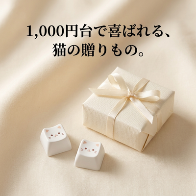リボン付きギフトボックスと白猫キーキャップ2つ。「1,000円台で喜ばれる、猫の贈りもの。」（価格はモック表現）