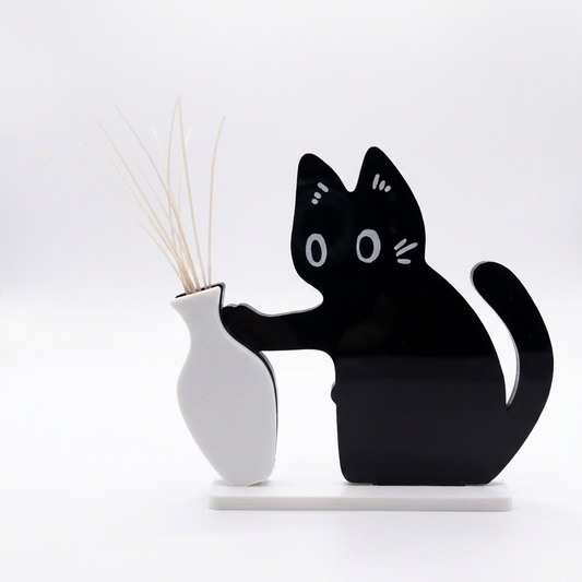 白い背景に置かれた、猫の形をした平らな黒いひげ飾りと白い一輪挿し。ひげが扇のように立っている。