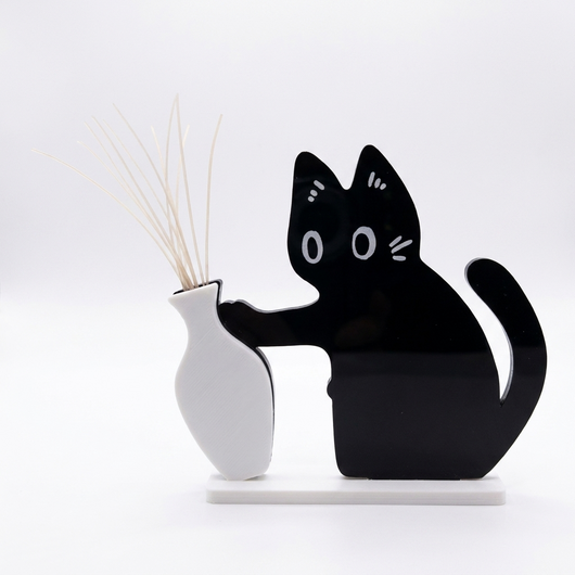 白い背景に置かれた、猫の形をした平らな黒いひげ飾りと白い一輪挿し。ひげが扇のように立っている。